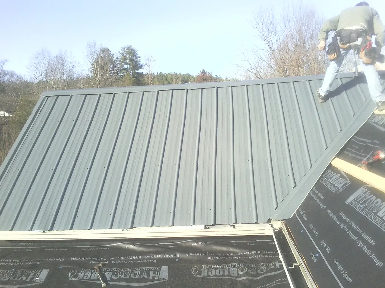 Crew installing dark green standing seam panels over synthetic underlayment in Glen Spey