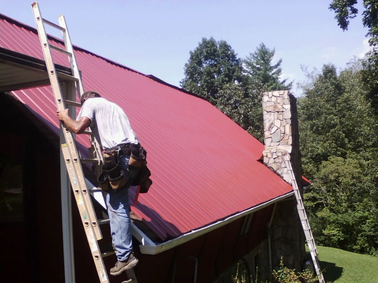 Roofer on a ladder beside a red metal roof and stone chimney — Soffit & Fascia Repair by Roofing Brothers in Glen Spey